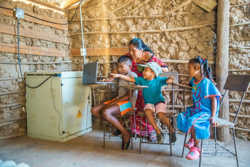 Mother and three children learning on laptops in a rustic mud-walled room.