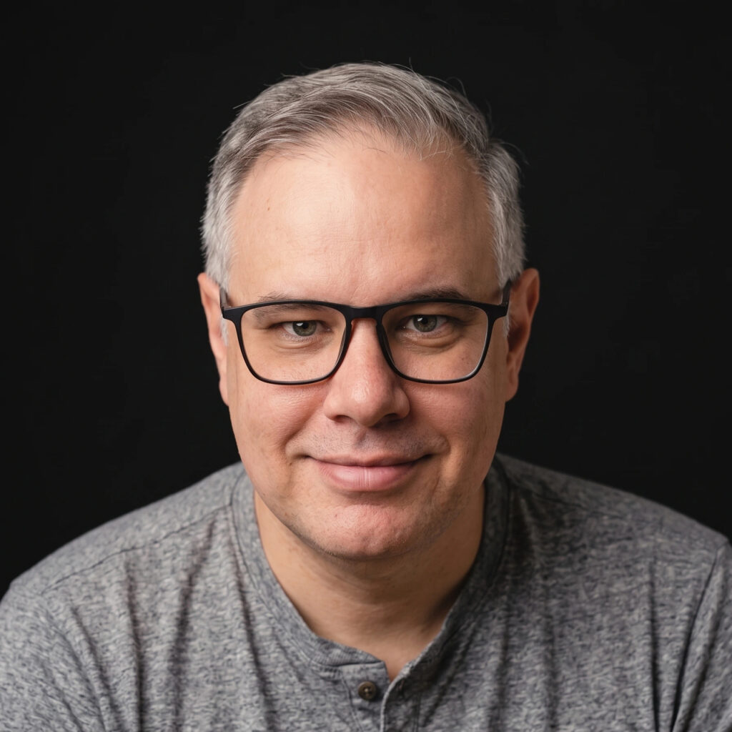 Close-up portrait of a middle-aged man with gray hair and black-ramed glasses, wearing a heather gray shirt against a black background.