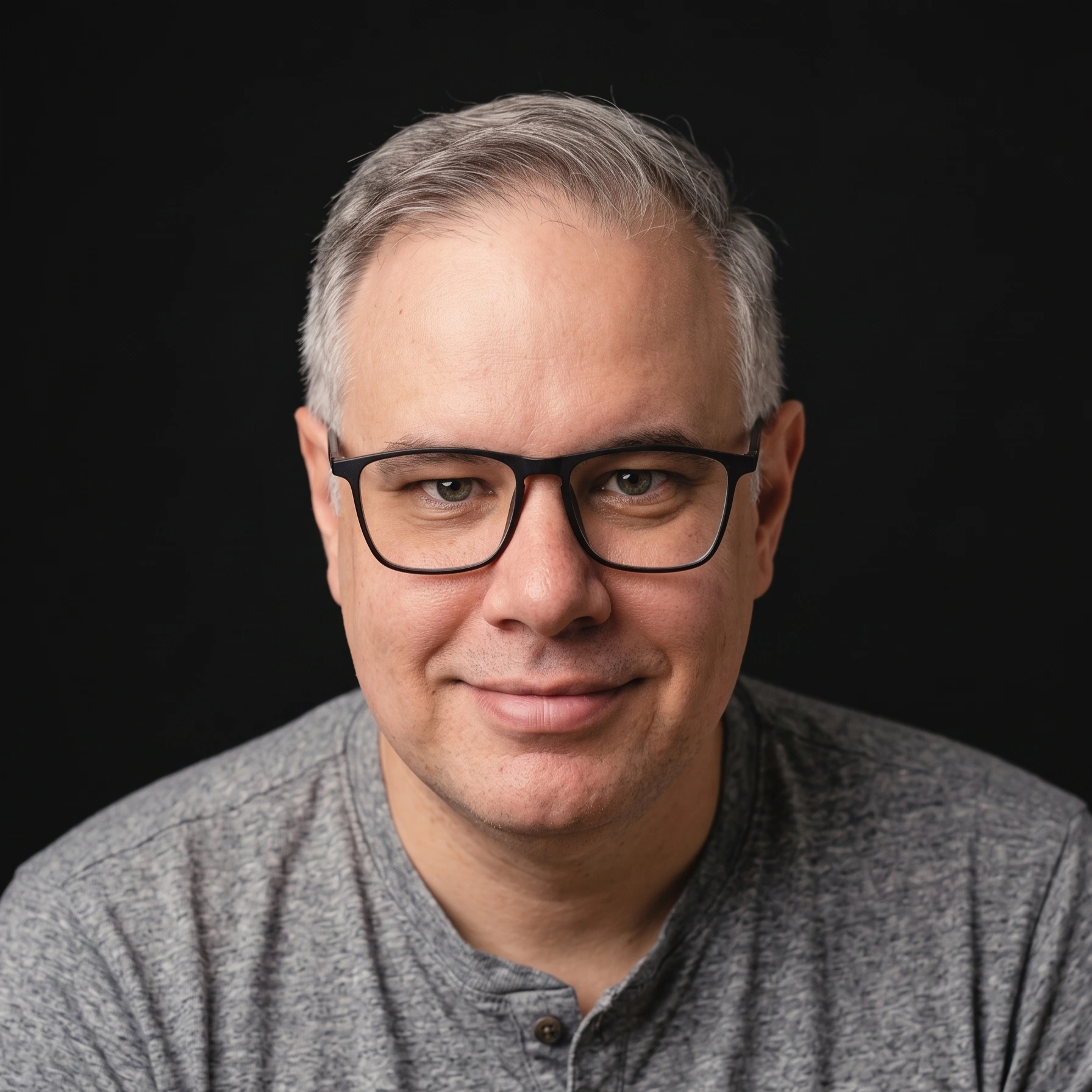 Close-up portrait of a middle-aged man with gray hair and black-ramed glasses, wearing a heather gray shirt against a black background.